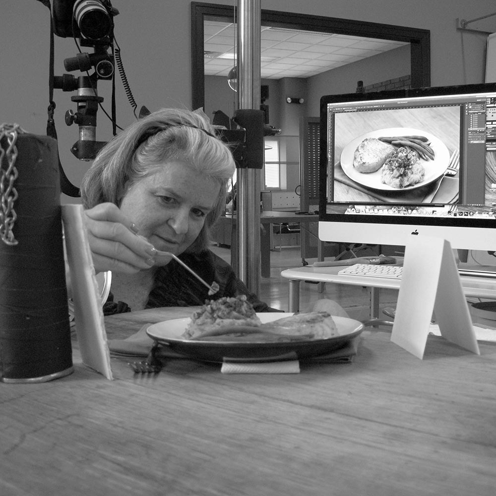 Black & white photo of Katy Keck styling with tweezers in a studio with camera gear and monitor of dish