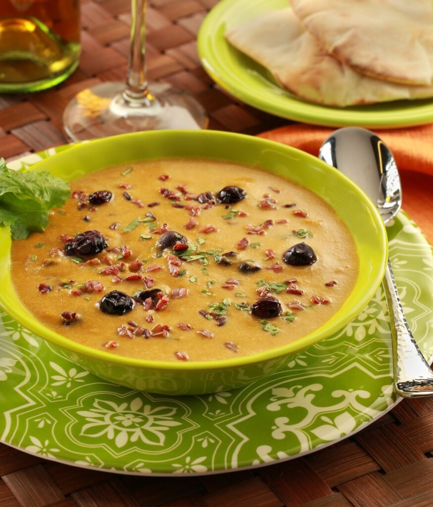 Mulligatawny soup with roasted blueberries and red rice in a green bowl on a green patterned small plate with a spoon, wine glass and small green plate with two pita breads