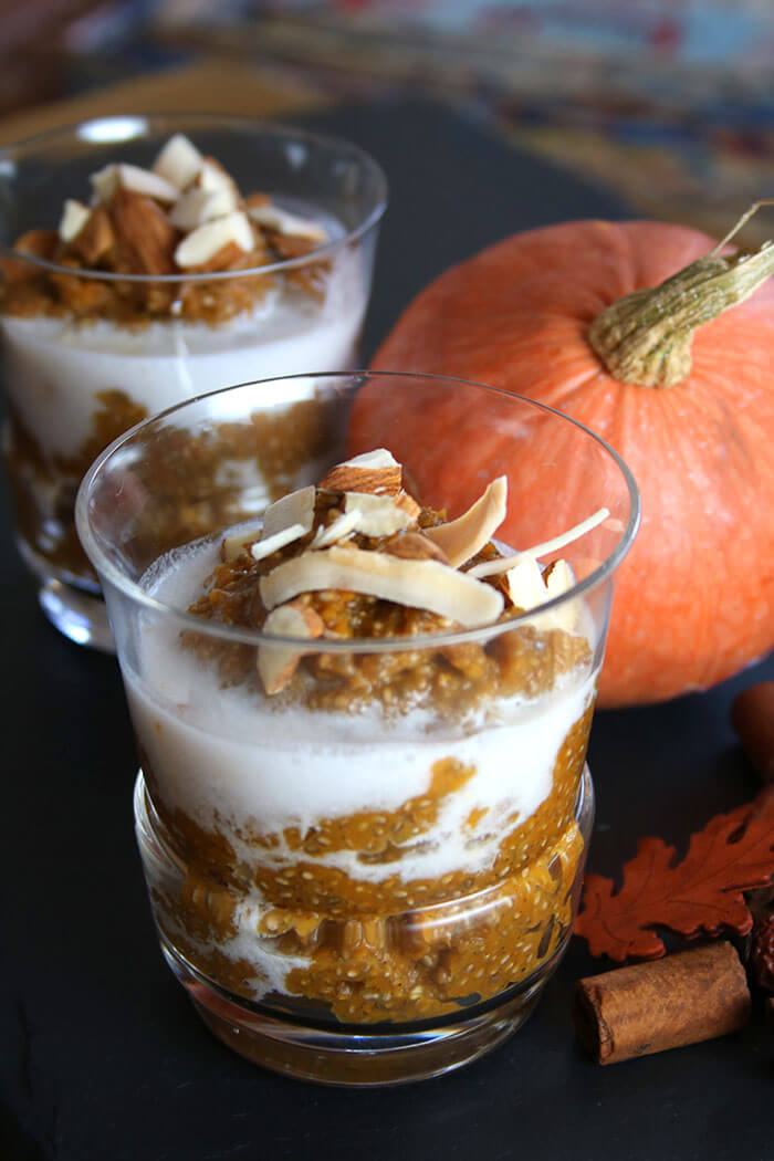 Pumpkin Chia Pudding in glasses topped with coconut milk, coconut strips, and almonds with a pumpkin behind