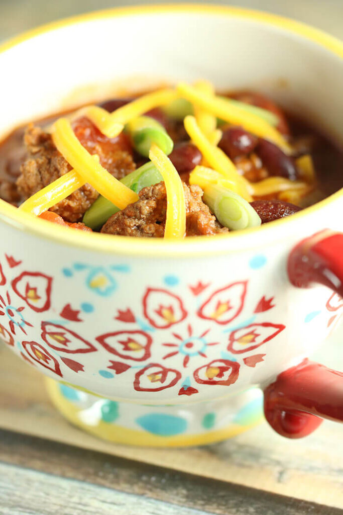 Mug with yellow rim and blue and red pattern, filled with chili topped with scallions and grated cheddar