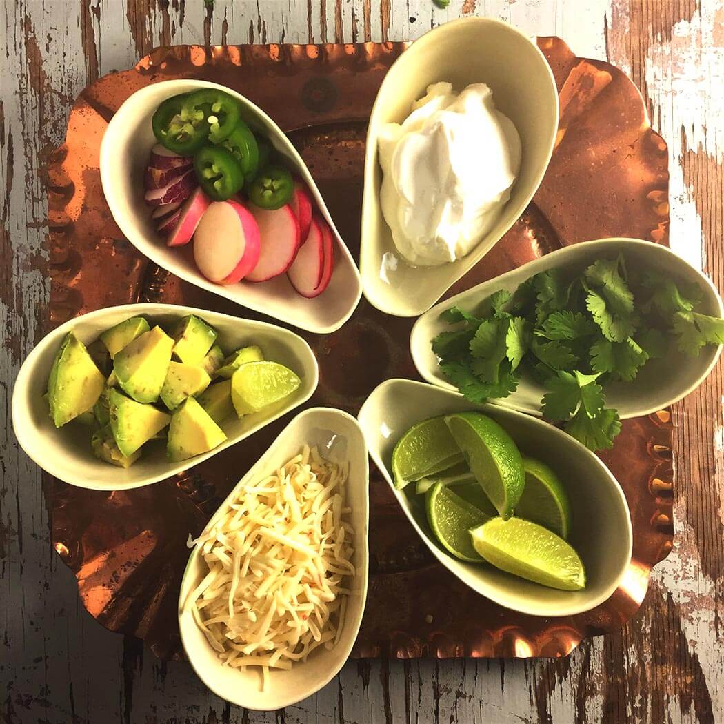overhead shot of Posole Toppings in bowls on a square copper tray: radish and jalapeno slices, sour cream, avocado chunks, grated monterey jack, lime wedges and cilantro sprigs