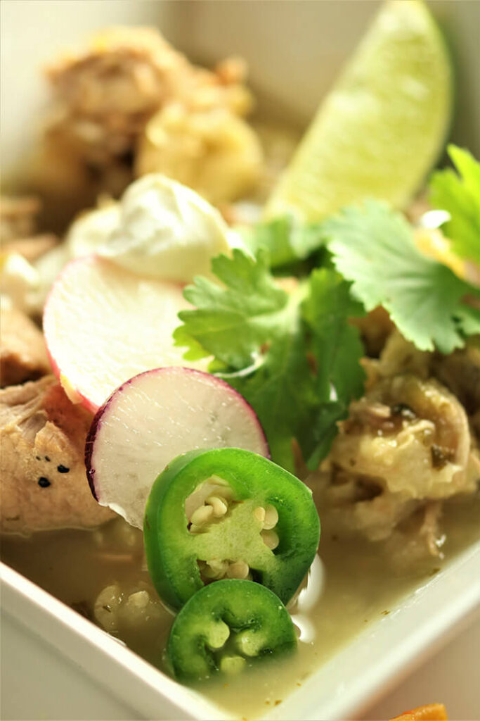 Posole with all the Fixin\'s in a square white bowl, topped with radish, jalapeno, sour cream and lime wedge