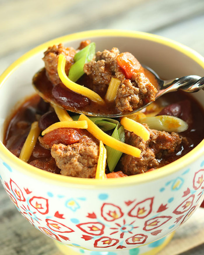 Mug with yellow rim and red and blue pattern, filled with chili and a spoon close up of chili with scallions and cheddar