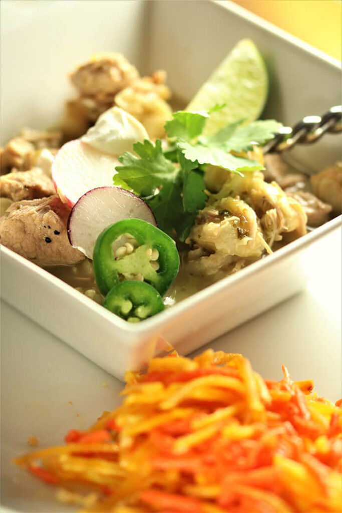 Southwestern Pork & Posole in a square white bowl topped with lime, sour cream, cilantro, radish slices and jalapenos, and served with carrot and yellow beet slaw