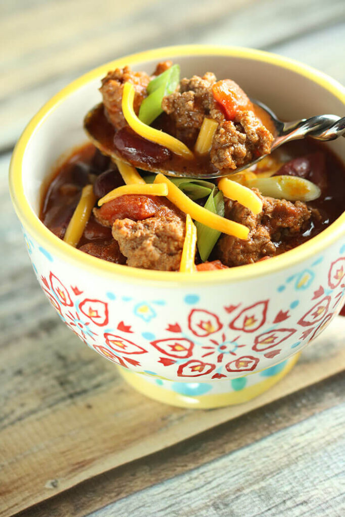 Mug with yellow rim and red and blue pattern, filled with chili and a spoon close up of chili with scallions and cheddar