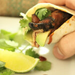 Hand holding a fig marinated chicken fajita with avocado, garnished with lime and cilantro