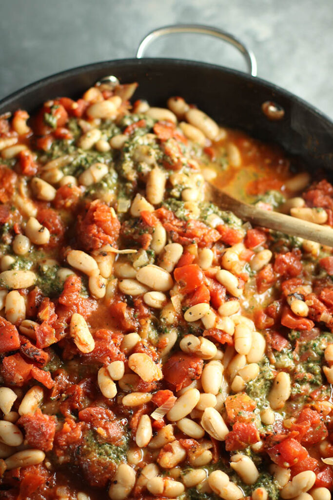 Large saute pan with a wooden spoon, tomatoes, pesto and white beans