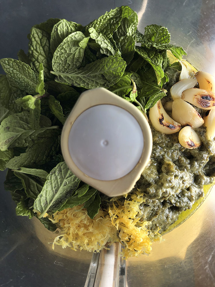 over head shot of a food processor work bowl with mint, pesto, roast garlic and lemon zest