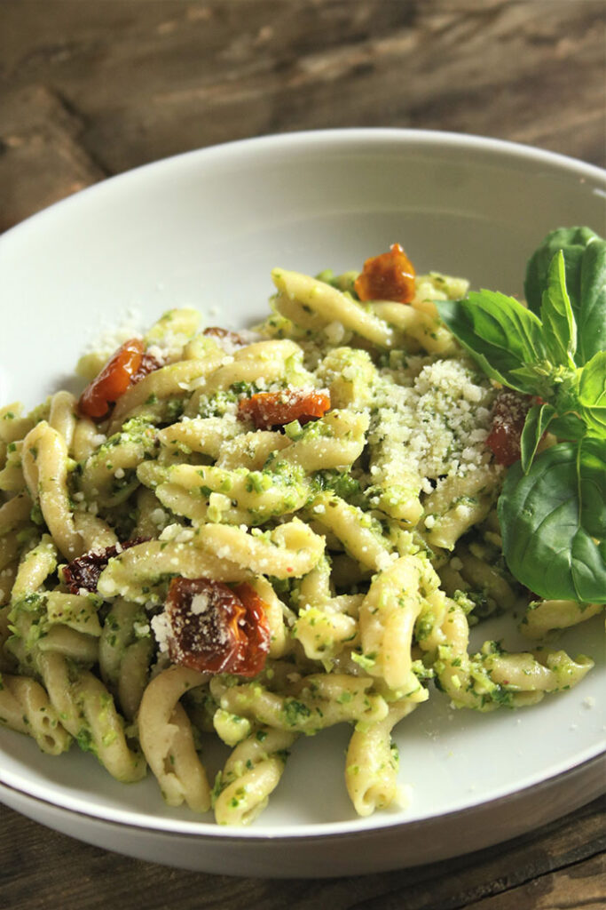 Scape Pesto on gemelli pasta with oven dried tomatoes in white bowl, garnished wit basil sprig