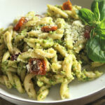 Scape Pesto on gemelli pasta with oven dried tomatoes in white bowl, garnished wit basil sprig