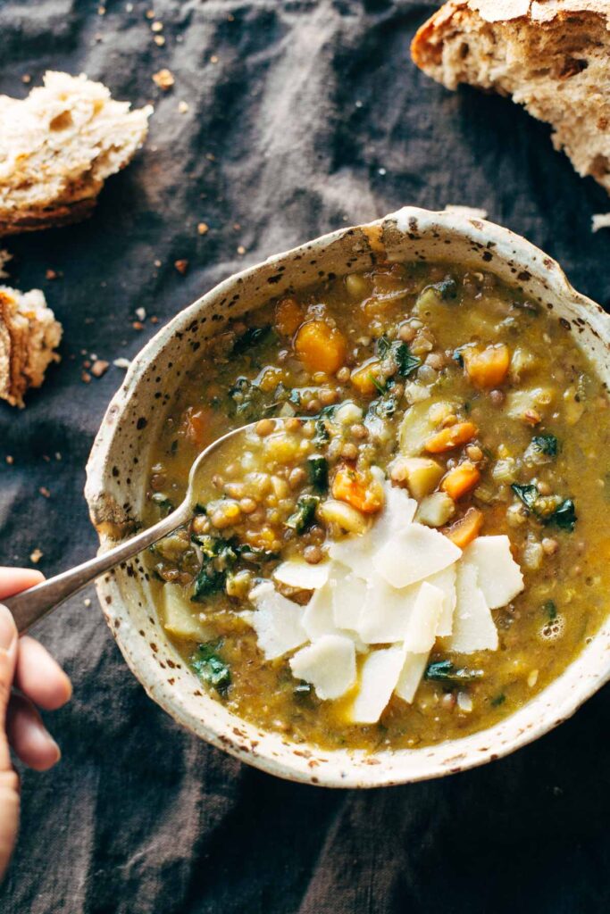 Pinch of Yum: crockpot lentil soup in a handmade pottery bowl with spoon. Soupd with greens and carrots and topped with parmesan shards. Served with bread