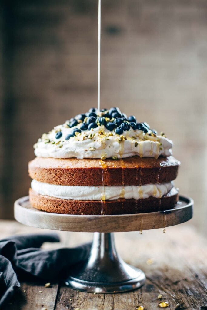 Pinch of Yum Orange brunch cake on a pedestal stand. Two layers of cake with whipped topping and filling and blueberries, honey being poured