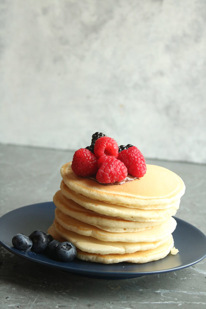 crooked stack of pancakes with raspberries on top and blue berries on the black plate