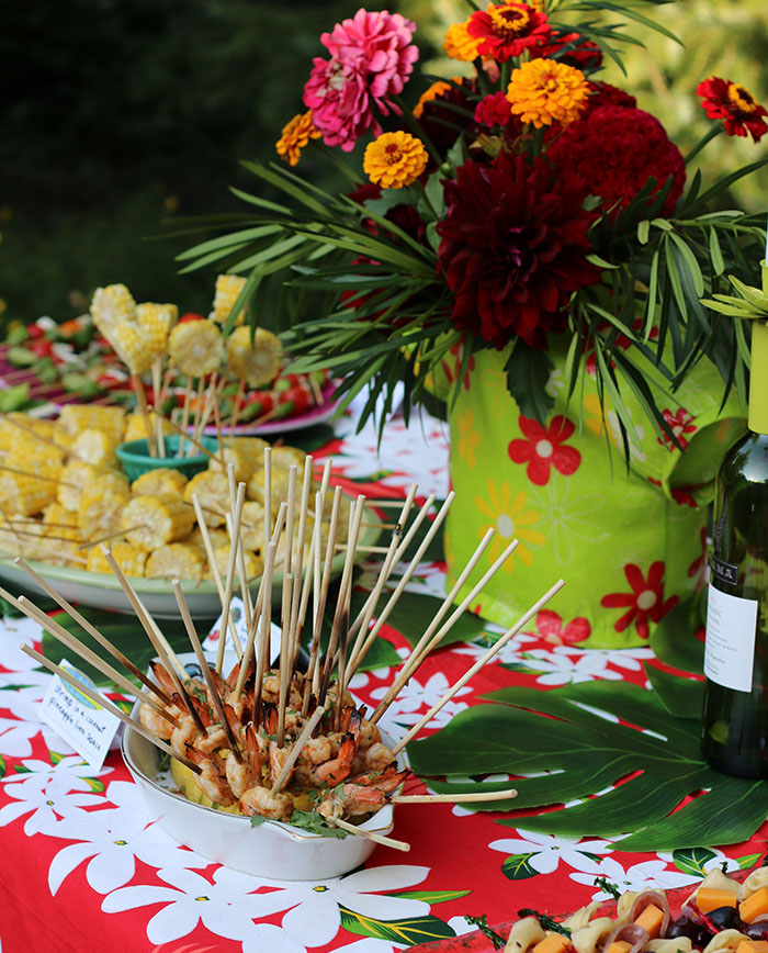 Food on a Stick: mini corn cobs, grilled shrimp and anti pasti skewers on a red cloth with white flowers and a Hawaiian shirt vase filled with flowers