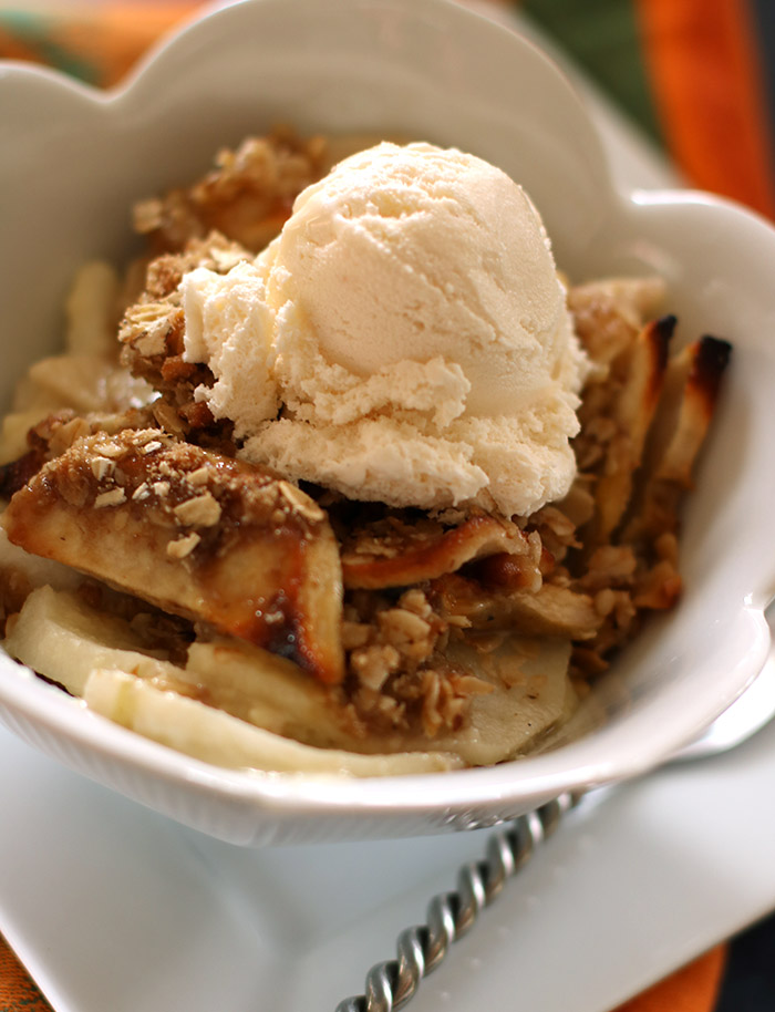 Apple crisp with vanilla ice cream in a scalloped edge bowl on a white square plate, twisted handle spoon underneath