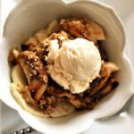 Apple crisp with vanilla ice cream in a scalloped edge bowl on a white square plate, twisted handle spoon underneath