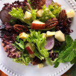 Harvest Salad overhead shot On a white plate with candied pecans, mixed greens, apple chunks and cheddar chunks, with purple radish slices
