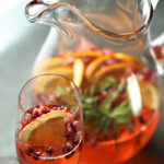 Glass of prosecco with orange slice and pomegranate arils, with a glass pitcher of the drink including rosemary sprigs