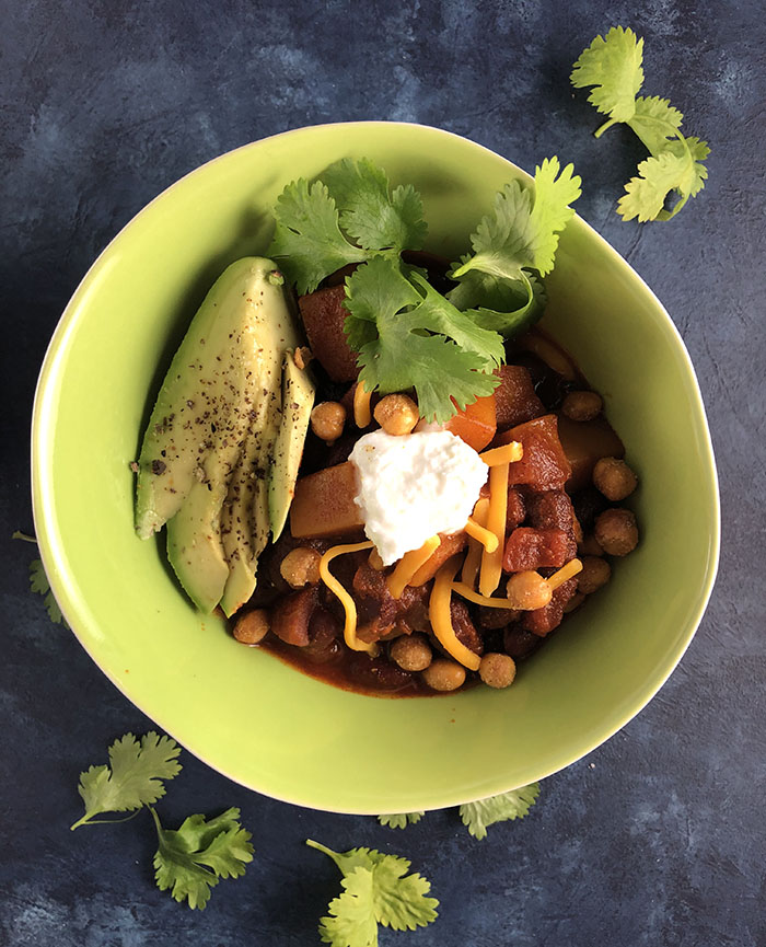 overhead view of butternut Squash Chili in a green bowl. Topped with avocado slices, sour cream, cheddar and crunchy garbanzos. Garnished with cilantro