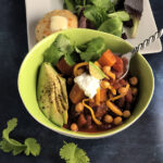 Butternut Squash Chili in a green bowl with a spoon. Topped with avocado slices, sour cream, cheddar and crunchy garbanzos. Garnished with cilantro and served with cheddar biscuit topped with butter and mixed green salad