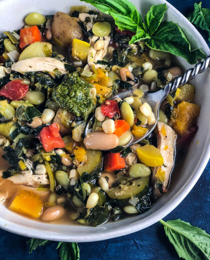 Hearty, Healthy Vegetable Soup with Chicken - White bowl with a spoon; includes white kidney beans, edamame, butternut squash, zucchini, leafy greens, small potatoes and pesto