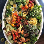 Southwestern Cobb: overhead view on a large white platter; deconstructed piles of avocado, corn, cherry tomatoes, hard boiled egg slices, limes, bacon, micro greens, grated jalapeno cheddar, grilled shrimp, salad greens and plums