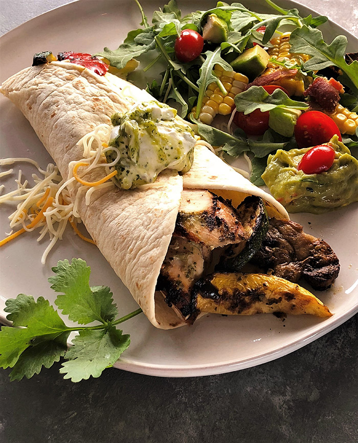 Salsa Verde Fajita: white plate with a flour tortilla, grilled vegetables and chicken, sauced with salsa verde and accompanied by an arugula, corn, tomato and avocado salad. Garnished with sour cream and cilantro