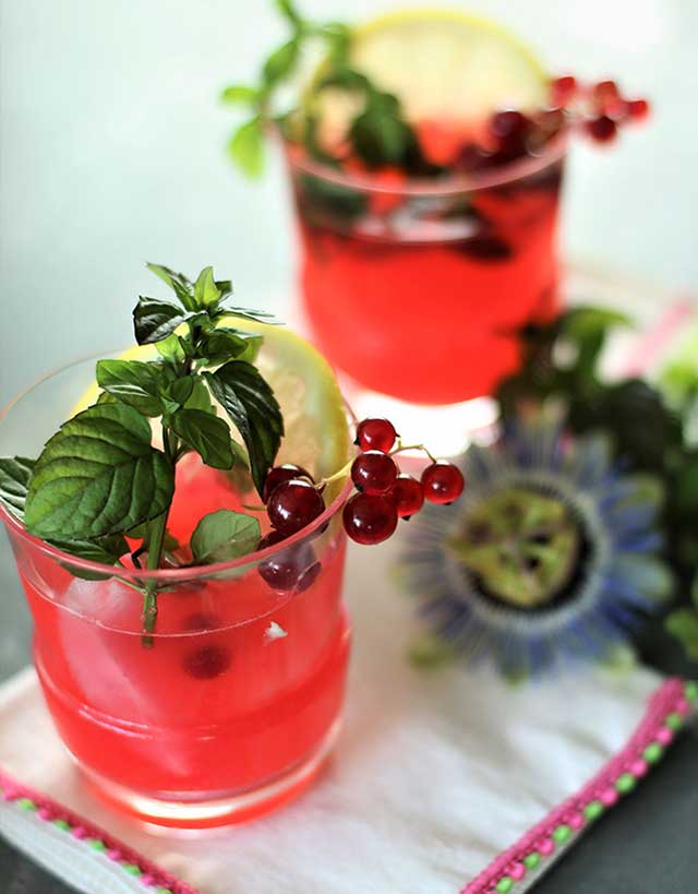 two glasses of red currant lemonaide with garnishes of currants, mint, lemon and a passion flower on the side