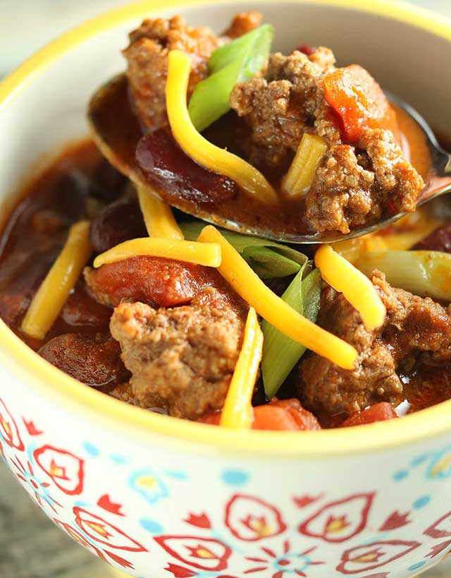 Bowl of cutty sark chili with spoon out, loaded with ground beef, scallions, tomatoes and grated cheddar