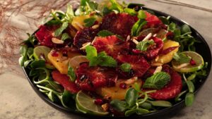 Black plate with bed of sunflower sprouts, topped with sliced navel oranges, blood oranges, red grapefruit and limes. Garnished with almonds, mint leaves, dates and pomegranate arils.