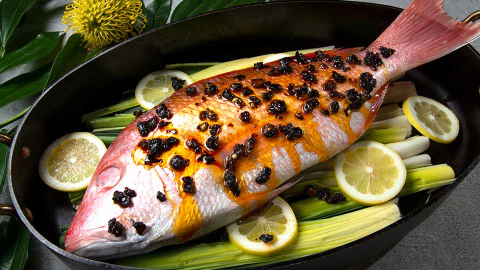 Whole red snapper with oo’mämē marinade in roasting pan on leeks and lemon slices.