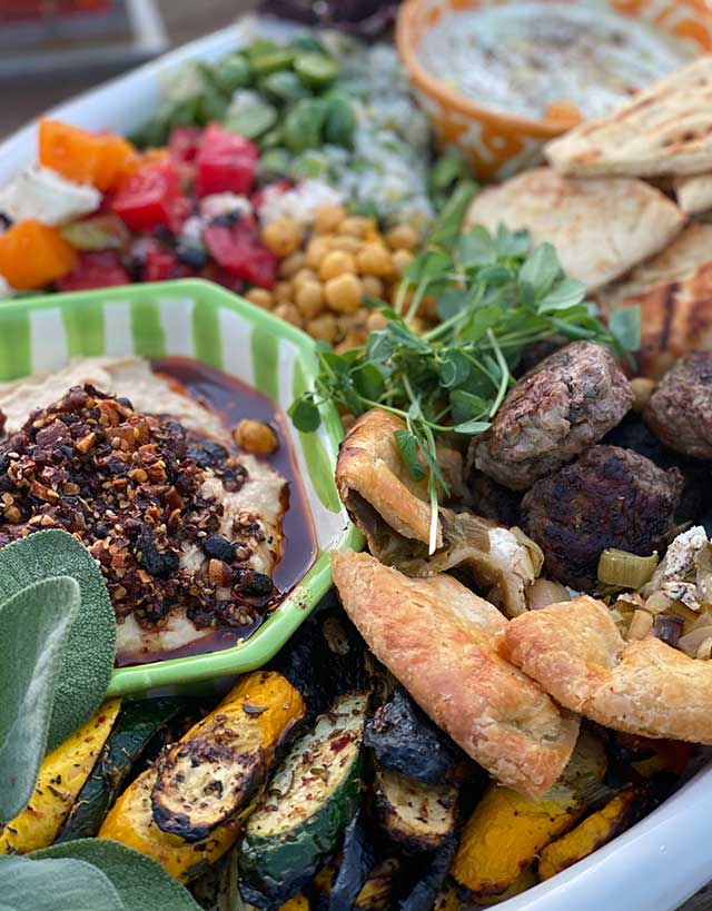 Large white oval platter with oo’mämē lamb sliders, leek galette, grilled squash, hummos topped with oo’mämē, tomato and sheep cheese, fonio with baby cucumbers, garbanzos, naan wedges, yogurt dip with spices and herb garnish.