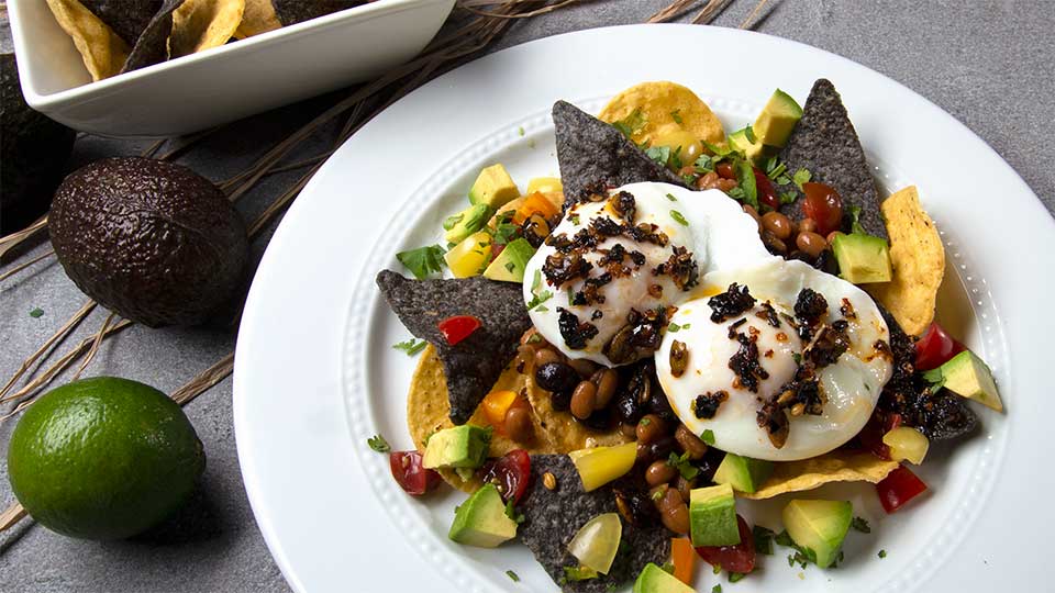 White round plate with chips, poached eggs, topped with Mexican oo’mämē Chile Crisp, avocado and tomatoes. White square bowl with chips and whole avocadoes and lime.