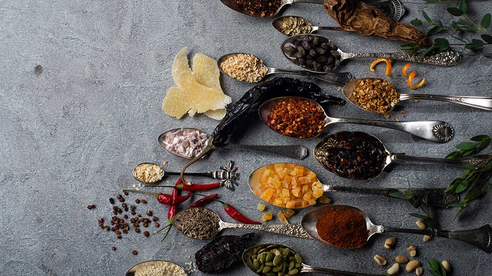 Vintage spoons filled with spices, seeds, chiles and fruits that are ingredients for oo’mämē chile crisp on slate grey background