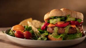 Veggie burger with a bun, green sauce and garnished with lettuce, carrots, tomatoes, cucumbers and sprouts. Garnished with cherry tomatoes and corn chips