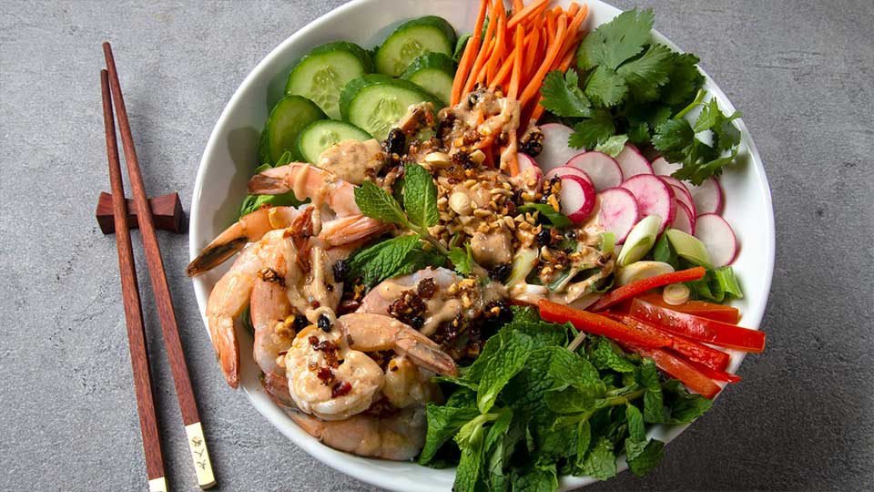 White shallow bowl with Chinese shrimp salad: Cucumbers, radishes, carrots, peppers, shrimp and herbs, topped with oo’mämē Chinese dressing
