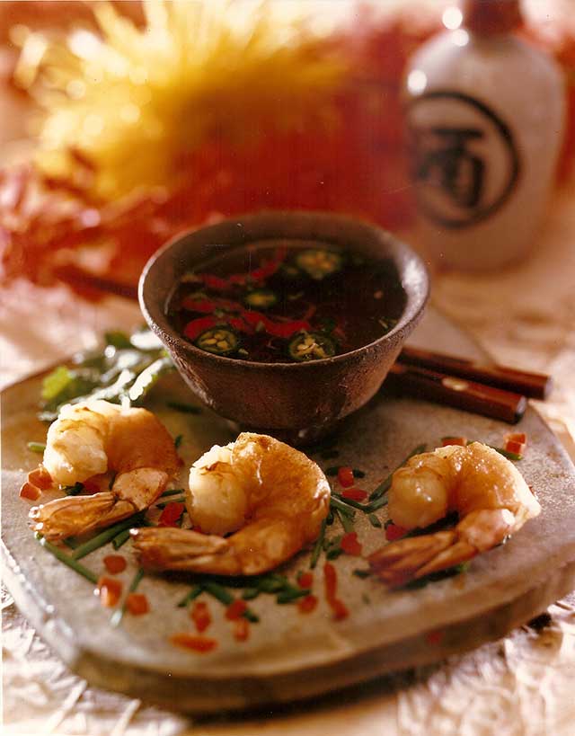 Three shrimp wrapped with wonts on a stone plate, with dipping sauce, chopsticks and flowers