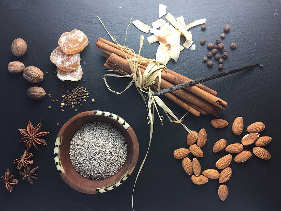 Overhead shot of black slate with nutmeg, star anise, crystallized ginger, cinnamon sticks, coconut flakes, vanilla bean and almonds