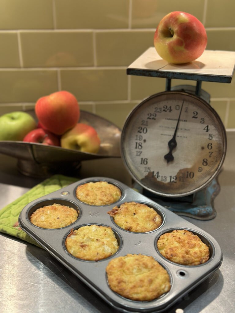 Corn muffins in a pan with a bowl of apples and an old time scale. 